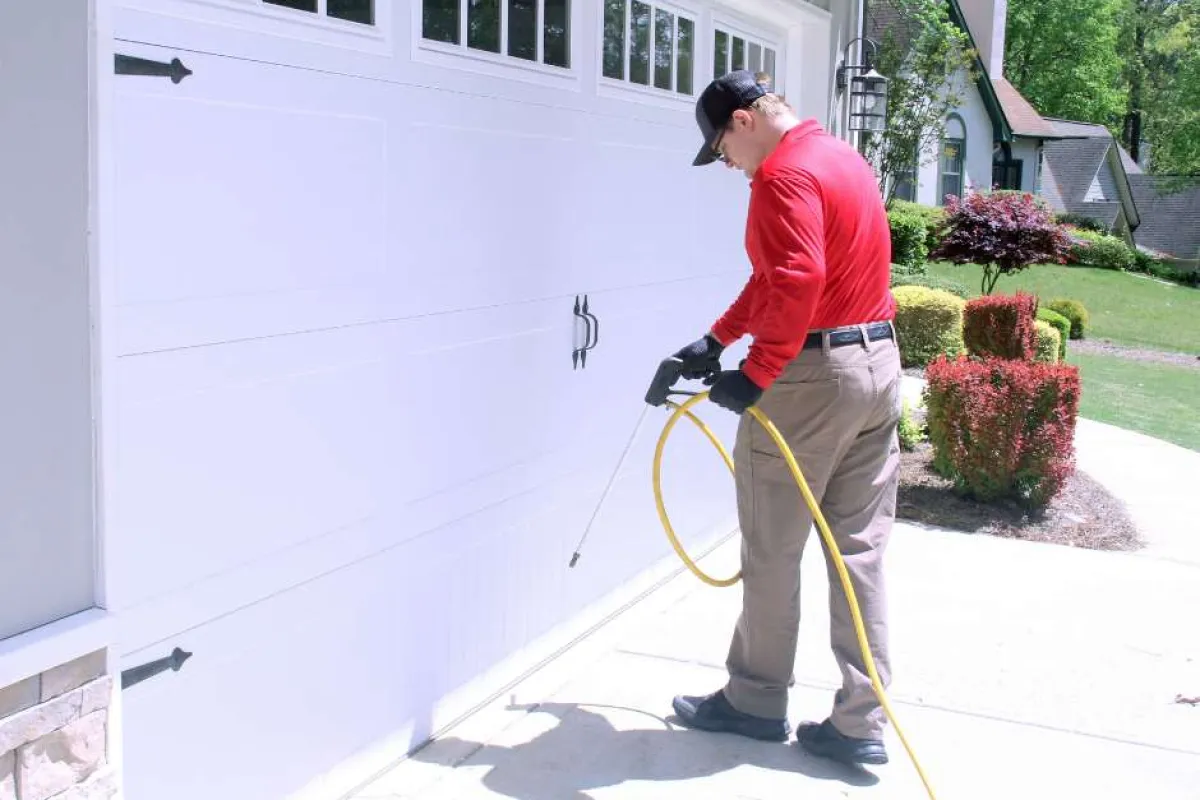 Man in red shirt spraying pesticide or cleaner on white garage door outdoors on sunny day