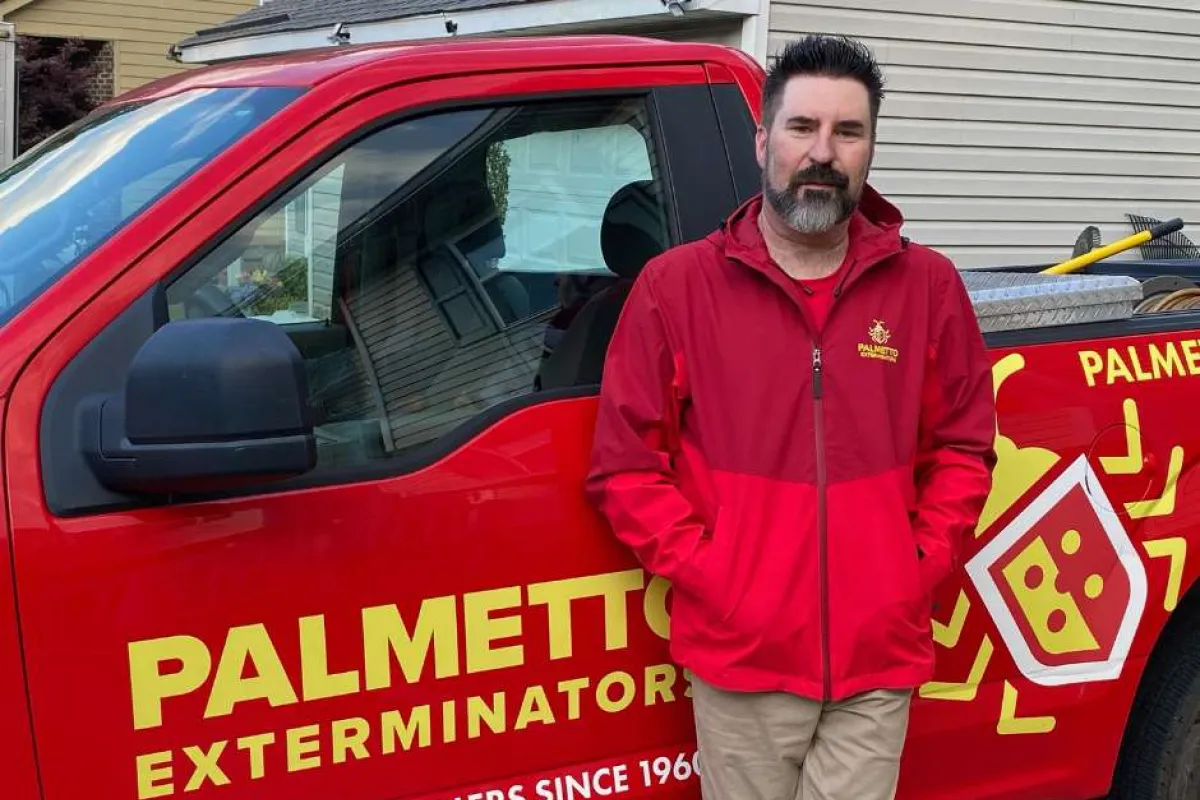 Man in red jacket stands next to red Palmetto Exterminators truck with yellow logo and text on side.