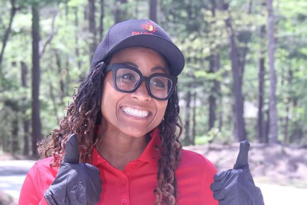 Smiling woman wearing a red shirt, black cap, black gloves, and large glasses giving two thumbs up outdoors.