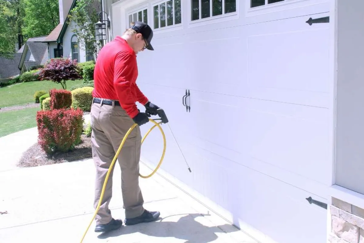 Man in red shirt spraying pesticide on garage door outdoors on a sunny day
