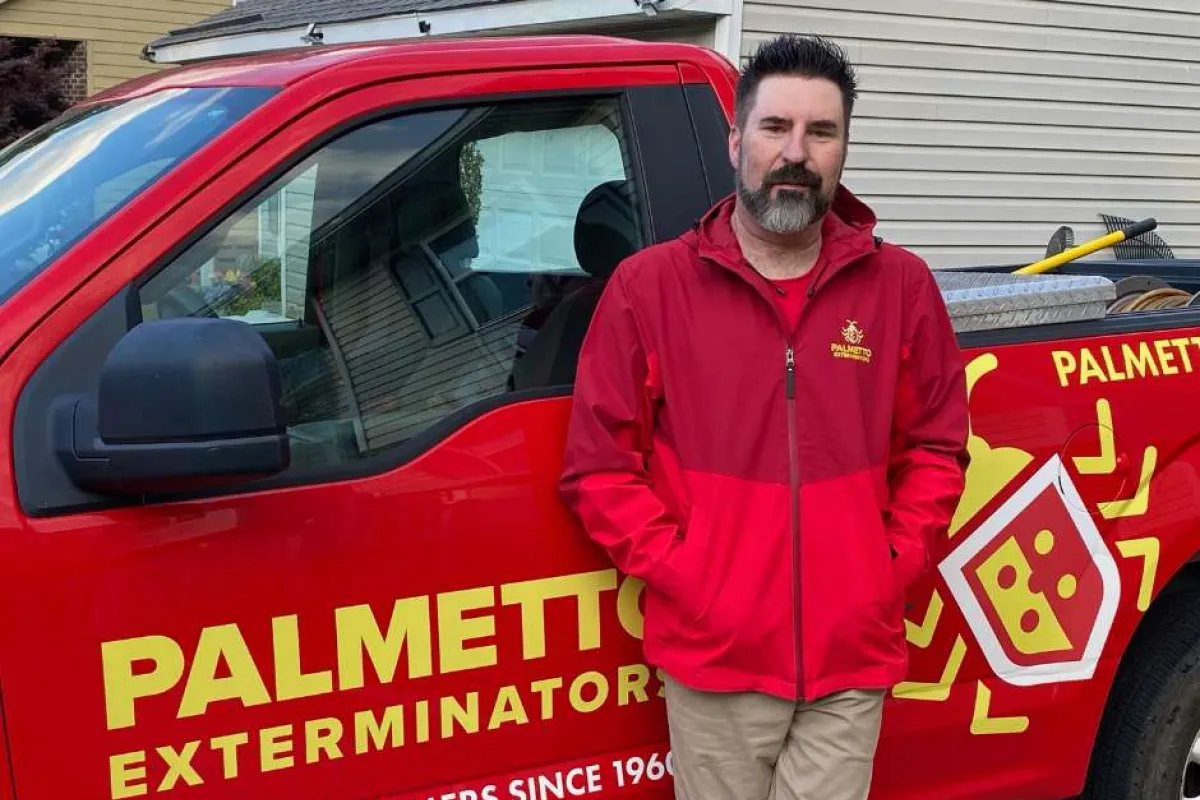 Man in red Palmetto Exterminators jacket leaning against branded red company truck with pest control logo.