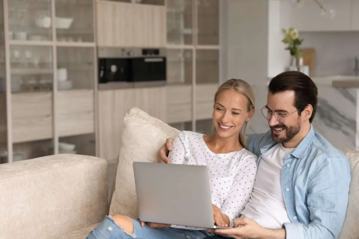 Happy couple sitting on couch using laptop together in a modern living room with kitchen background