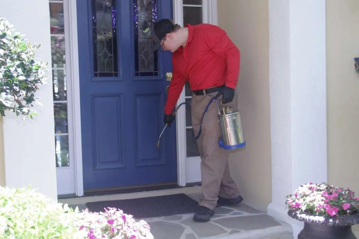 Pest control worker in red shirt spraying insecticide near blue front door of a house with flowers.