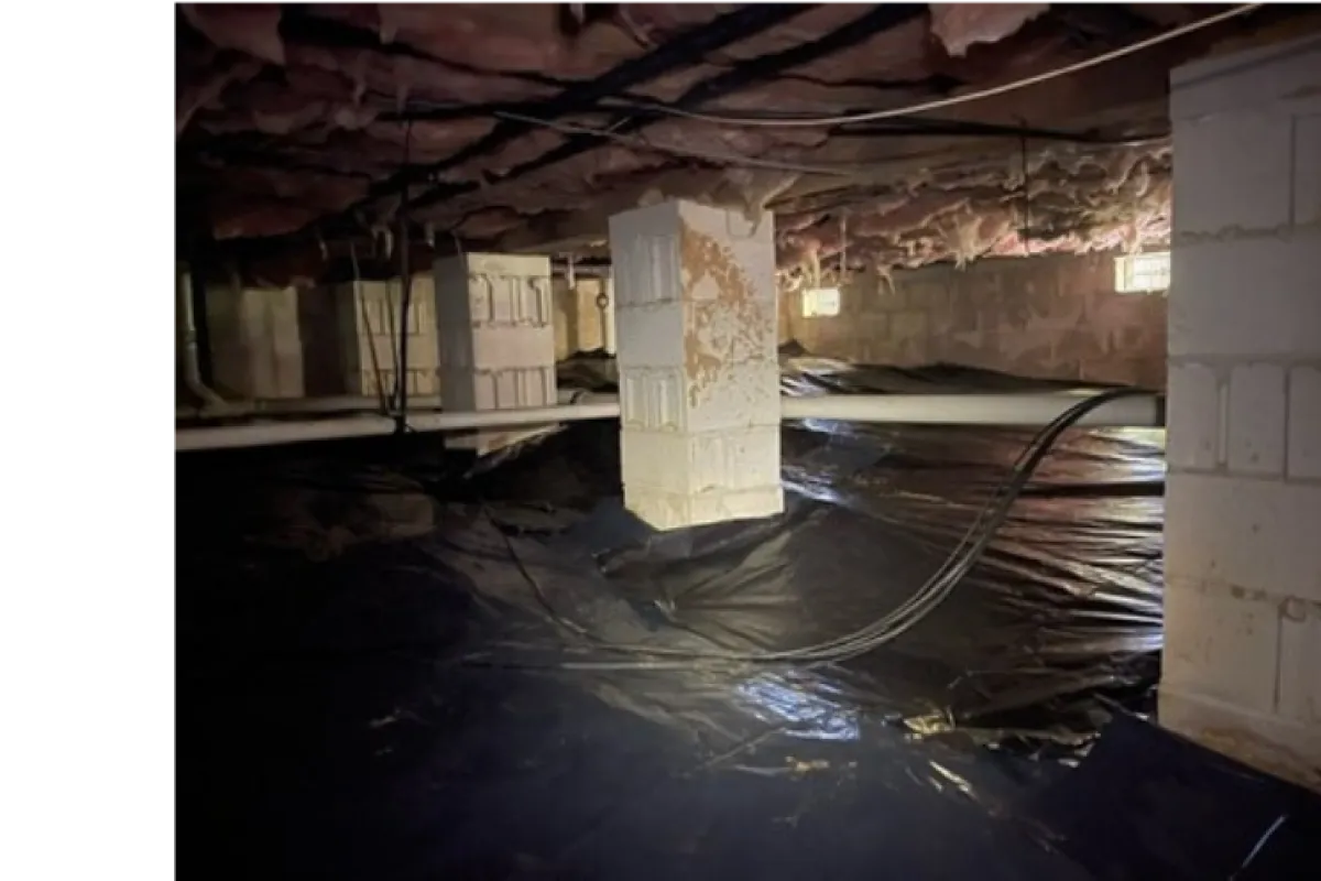 Dark crawl space with plastic vapor barrier covering dirt floor and support pillars under house ceiling insulation.