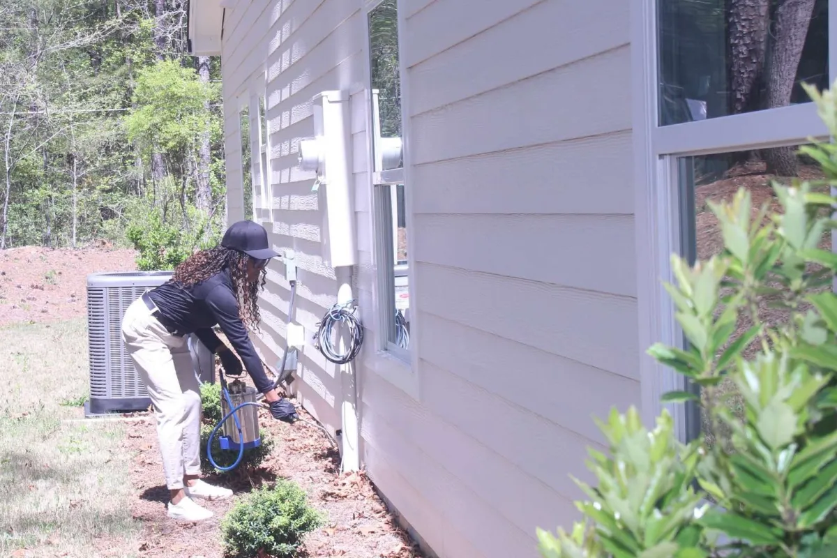 Woman spraying pesticide on plants along the side of a white house on a sunny day