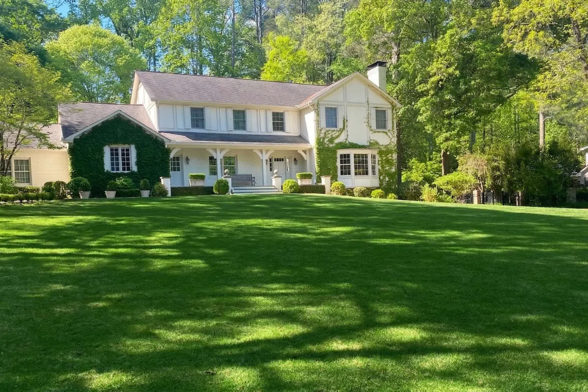 Two-story white house with ivy on facade surrounded by lush green lawn and tall trees in bright sunlight.