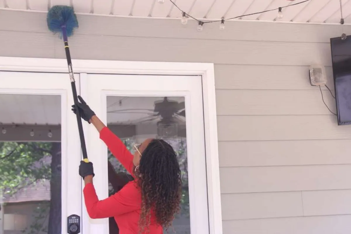Woman in red shirt dusting ceiling light fixture with an extendable duster outside a house.