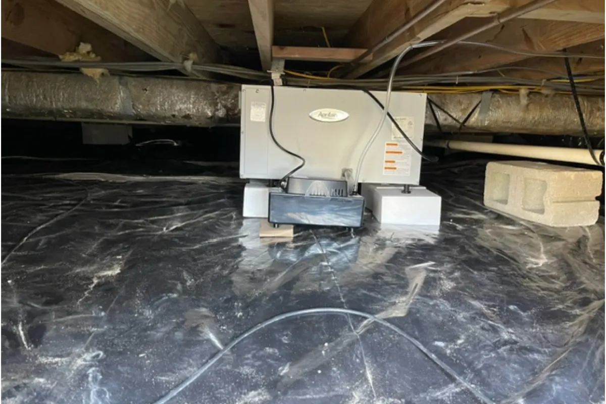 Dehumidifier installed in a crawl space with a black vapor barrier and exposed wooden beams above.
