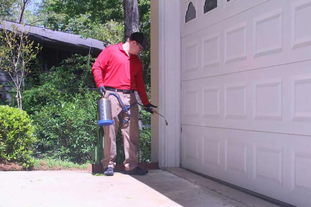 Man in red shirt applying pest control spray along the base of a residential garage door outdoors.