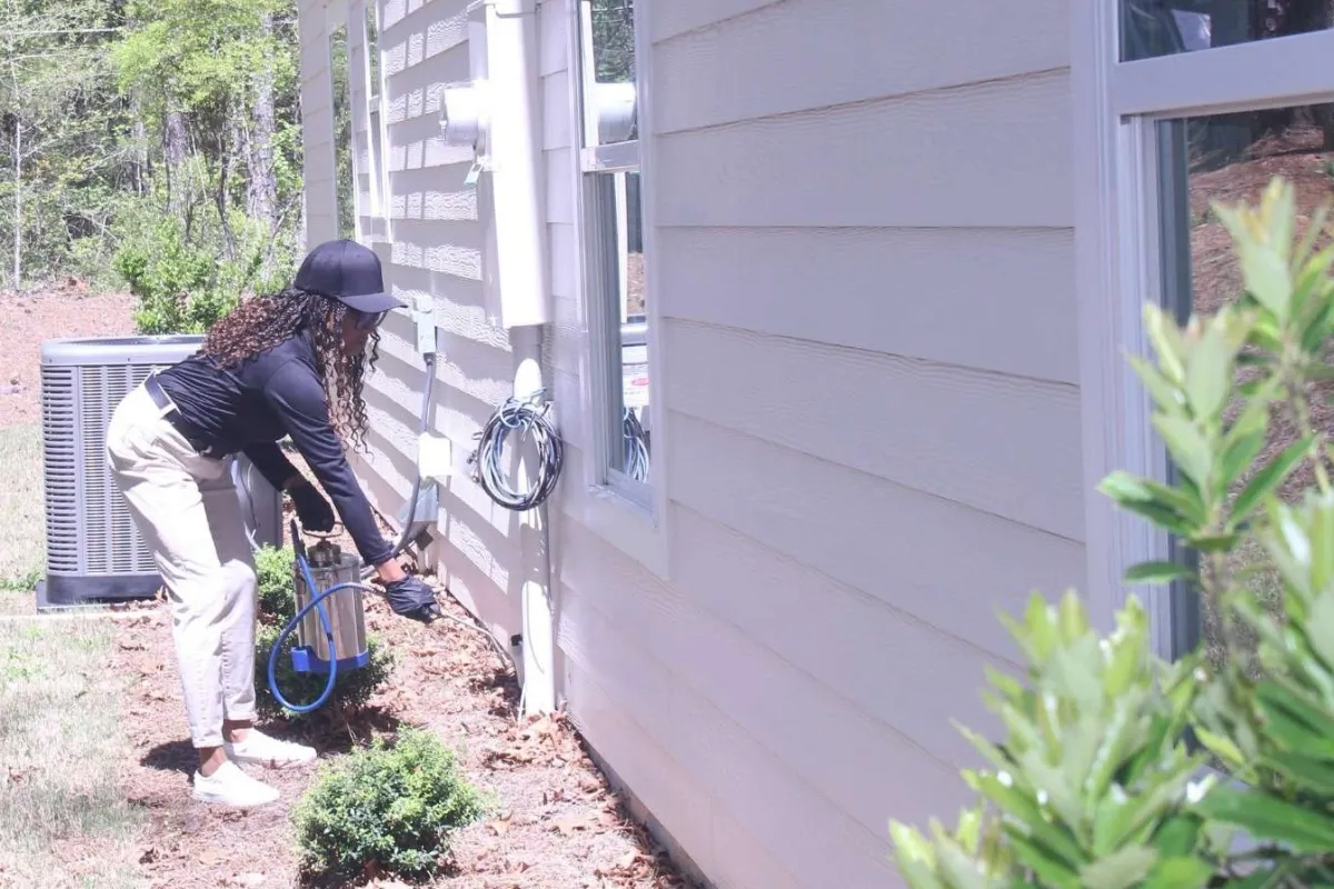 Person in gloves and cap spraying plants beside a white house with greenery and air conditioning unit outside.