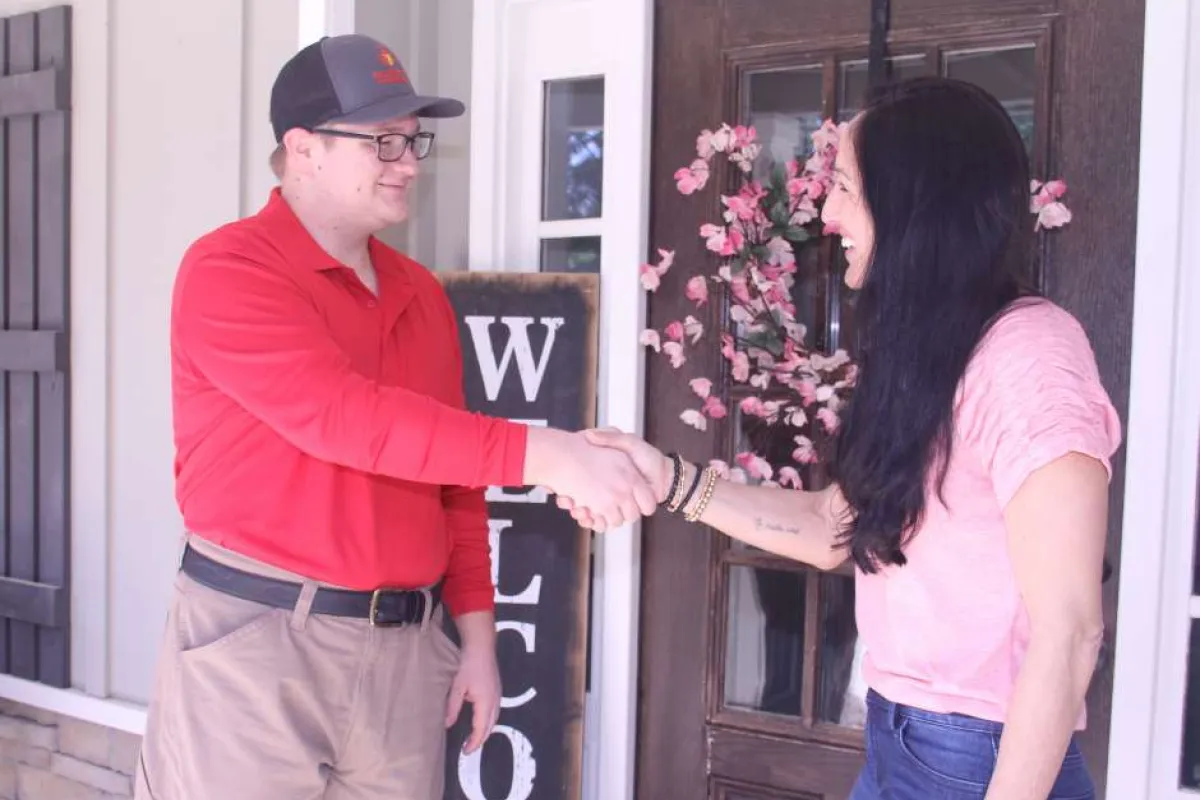 Man in red shirt shaking hands with woman in pink shirt at front door with welcome sign and flowers.