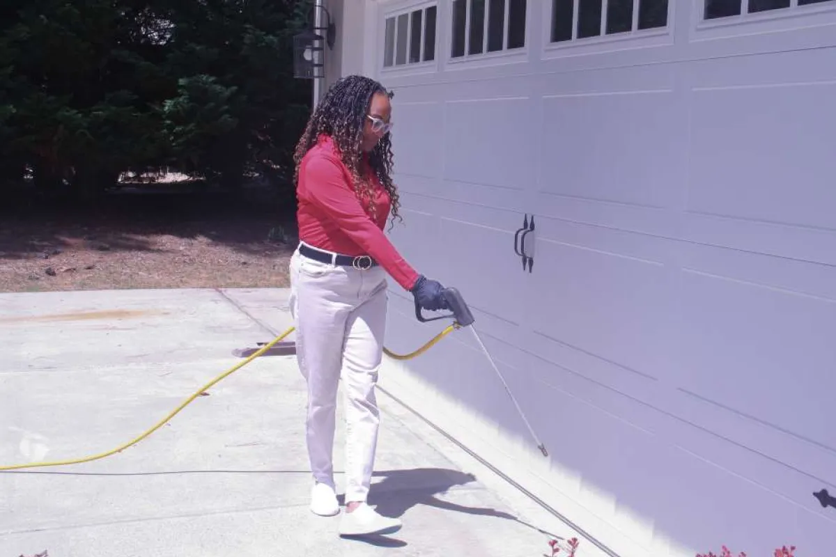Woman wearing gloves sprays pesticide around a garage door on a sunny day outdoors.
