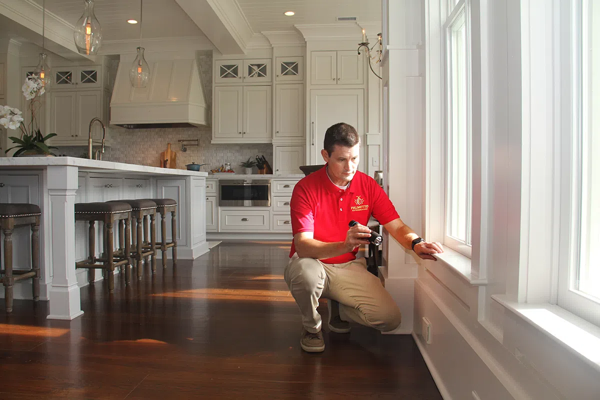 Man in red shirt inspecting white window frame in modern kitchen with hardwood floors and island seating.
