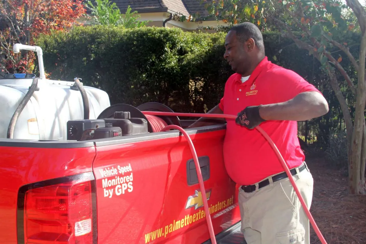 Pest control worker in red polo loading hose from red Palmetto Exterminators truck in a residential area.