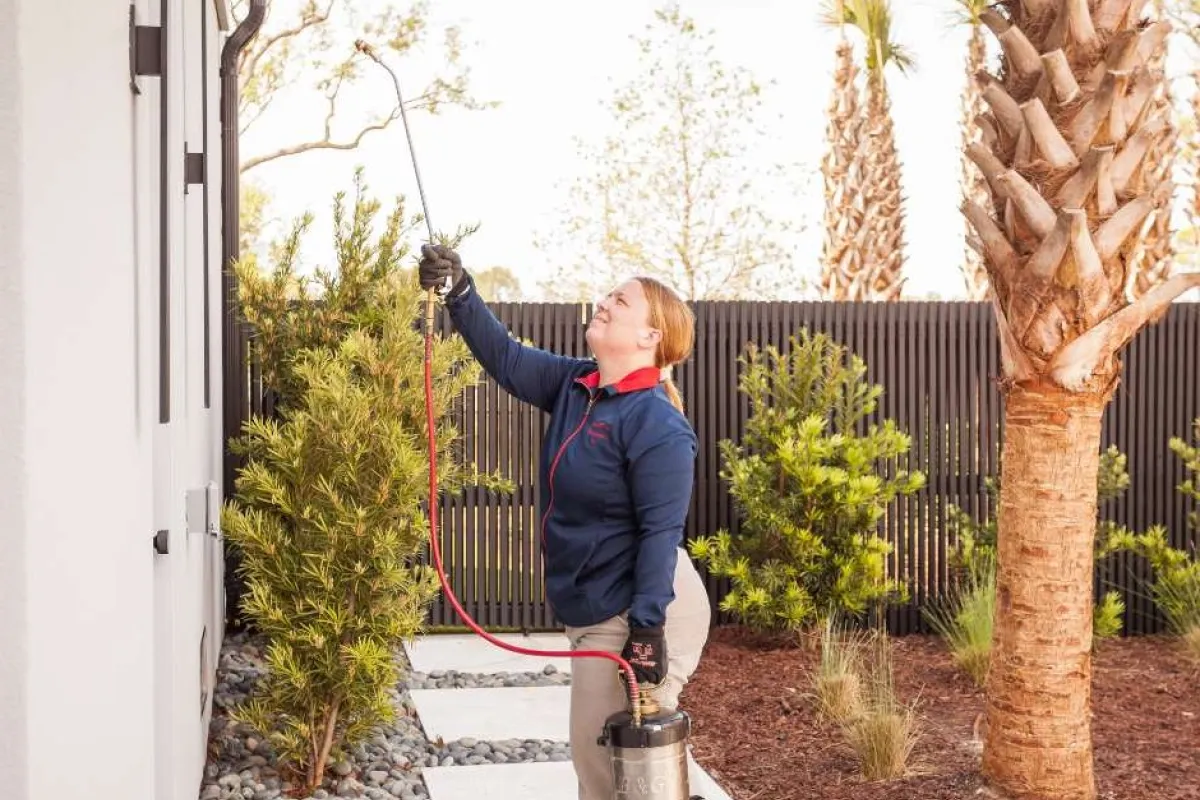 Pest control technician spraying exterior of home with pesticide using spray tank in landscaped yard.