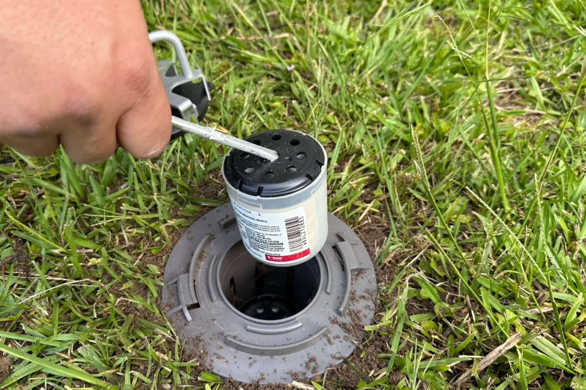 Hand using tool to remove sprinkler head cap from irrigation valve set in green grass lawn.