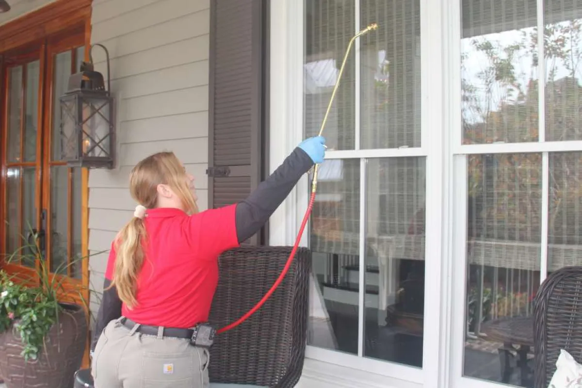Technician in red shirt applying pest control spray on house exterior window from wicker chair porch area.