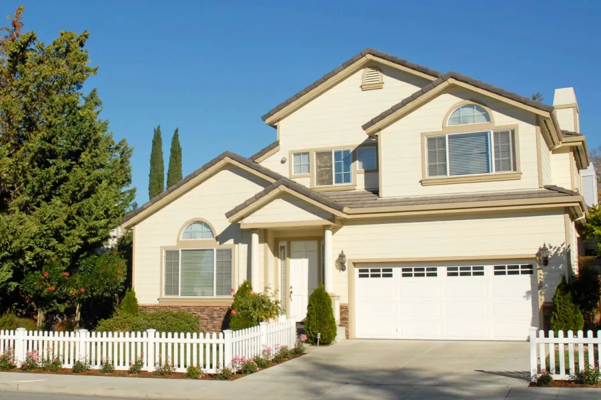Two-story beige house with white picket fence, large windows, garage, and landscaped front yard under clear blue sky