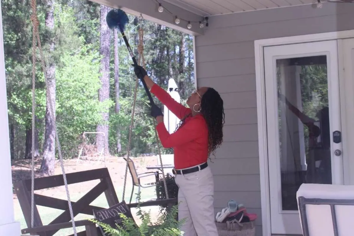 Woman cleaning porch ceiling with a long-handled duster on a sunny day with green trees outside.