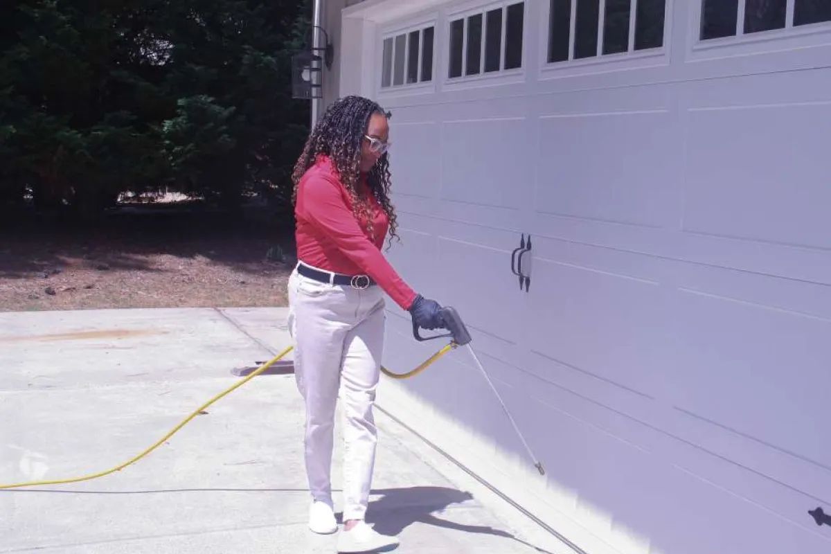 Woman wearing gloves spraying pesticide along the base of a garage door with a hose and spray wand.