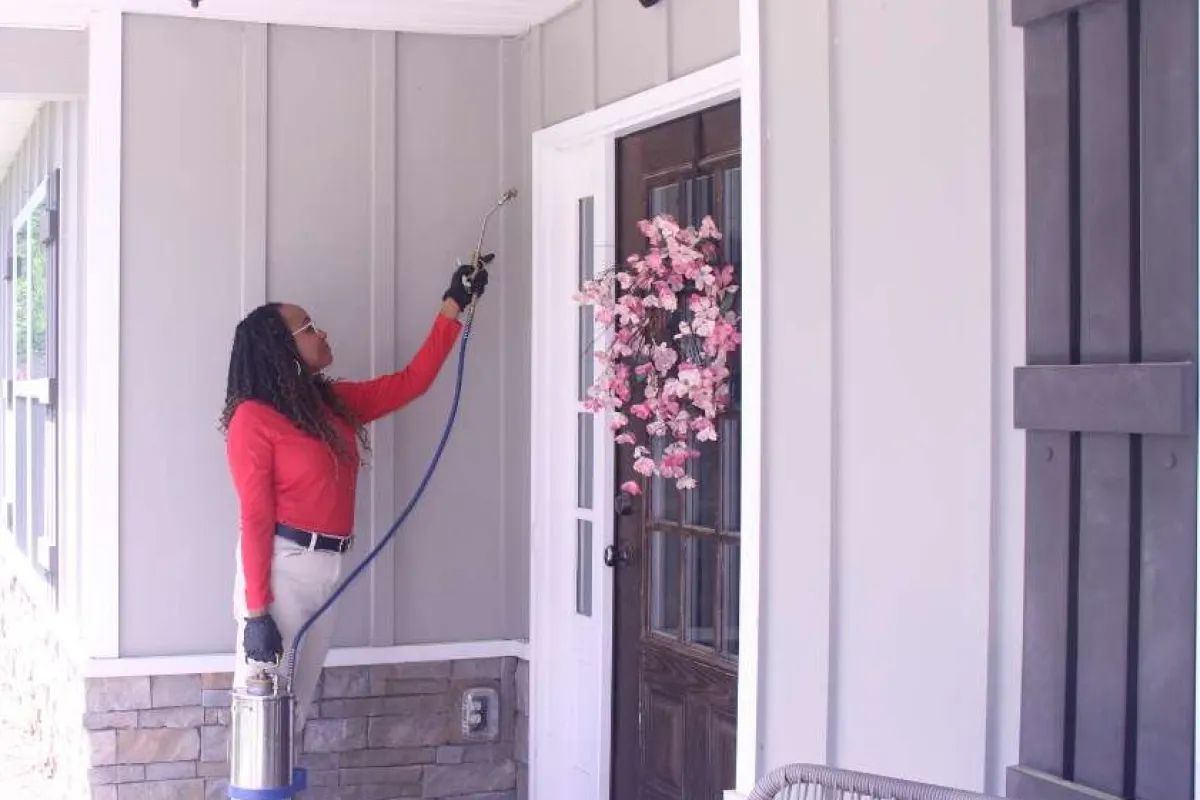 Woman applying pest control spray to house exterior near front door with flower wreath.