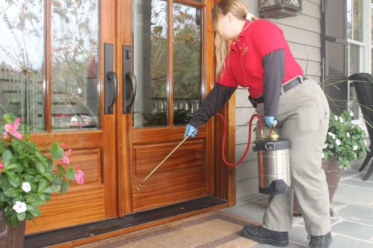 Pest control technician spraying insecticide at a wooden front door of a house with plants nearby