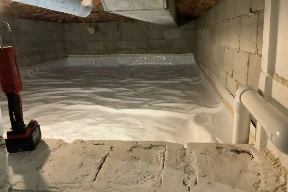 Crawl space with vapor barrier installed on ground, surrounded by concrete block walls and ventilation ducts.