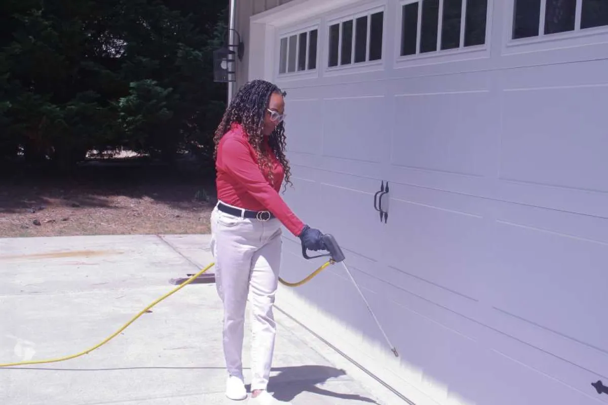 Woman wearing gloves spraying pesticide along the base of a garage door outdoors on a sunny day.