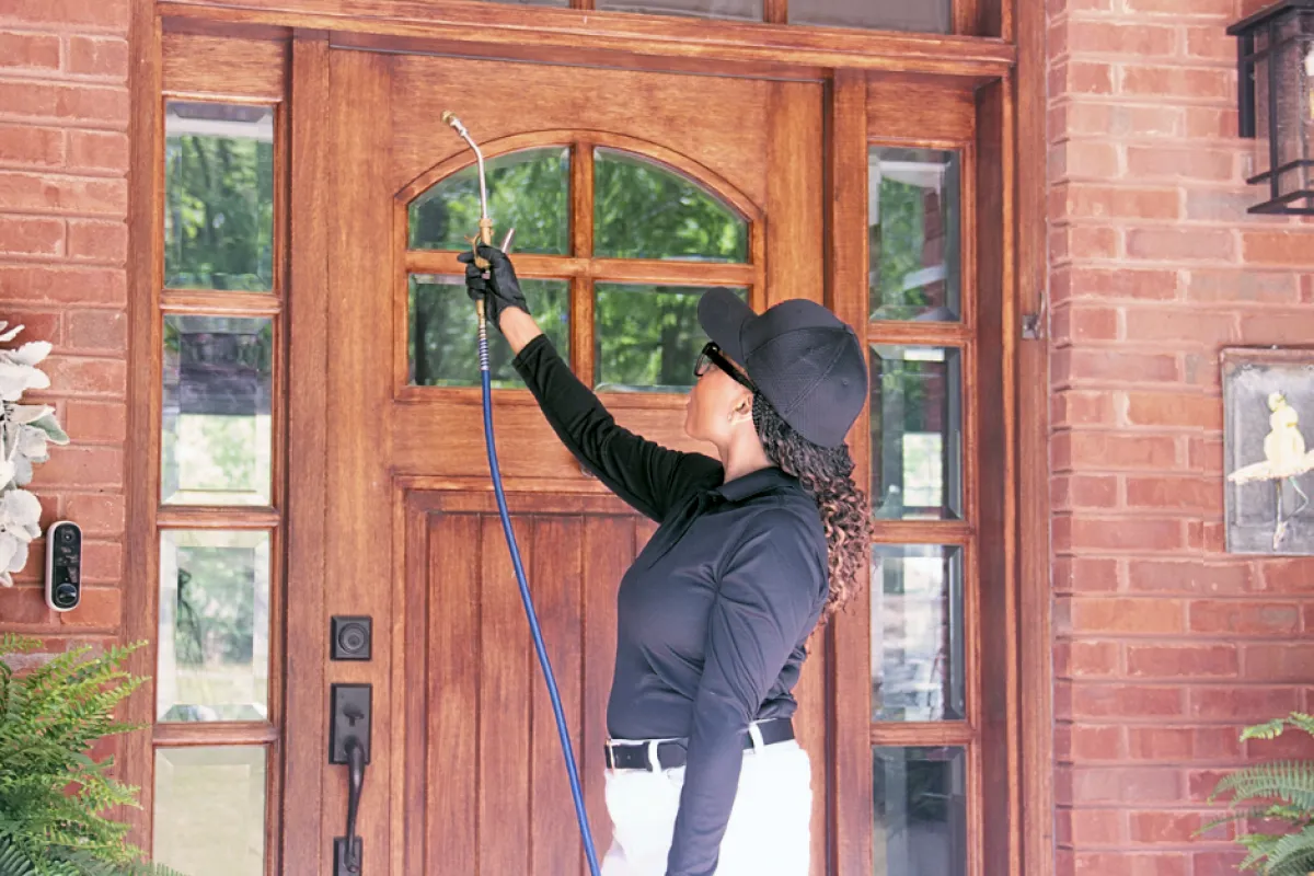 Technician wearing black cap spraying pesticide on a wooden front door with brick walls outside a house.