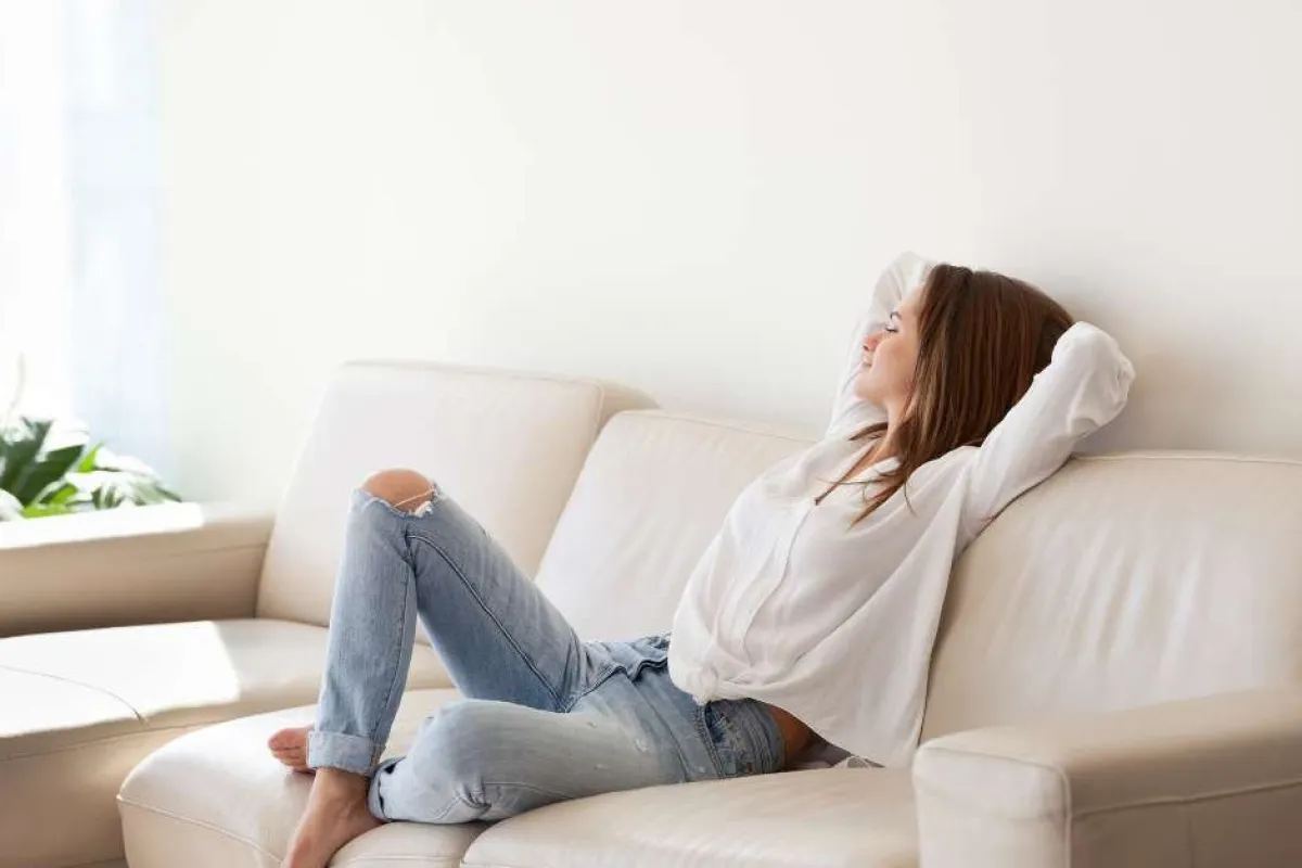 Young woman relaxing on a beige leather sofa in a bright, minimalist living room with natural light.