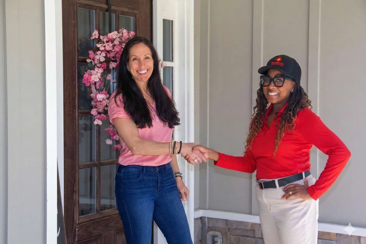 Two women shaking hands in front of a door with pink flowers, smiling and dressed casually in bright colors.