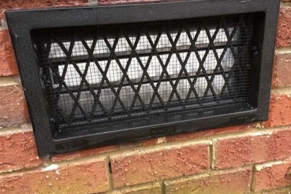 Black metal vent cover with diamond mesh pattern installed on a red brick wall exterior.