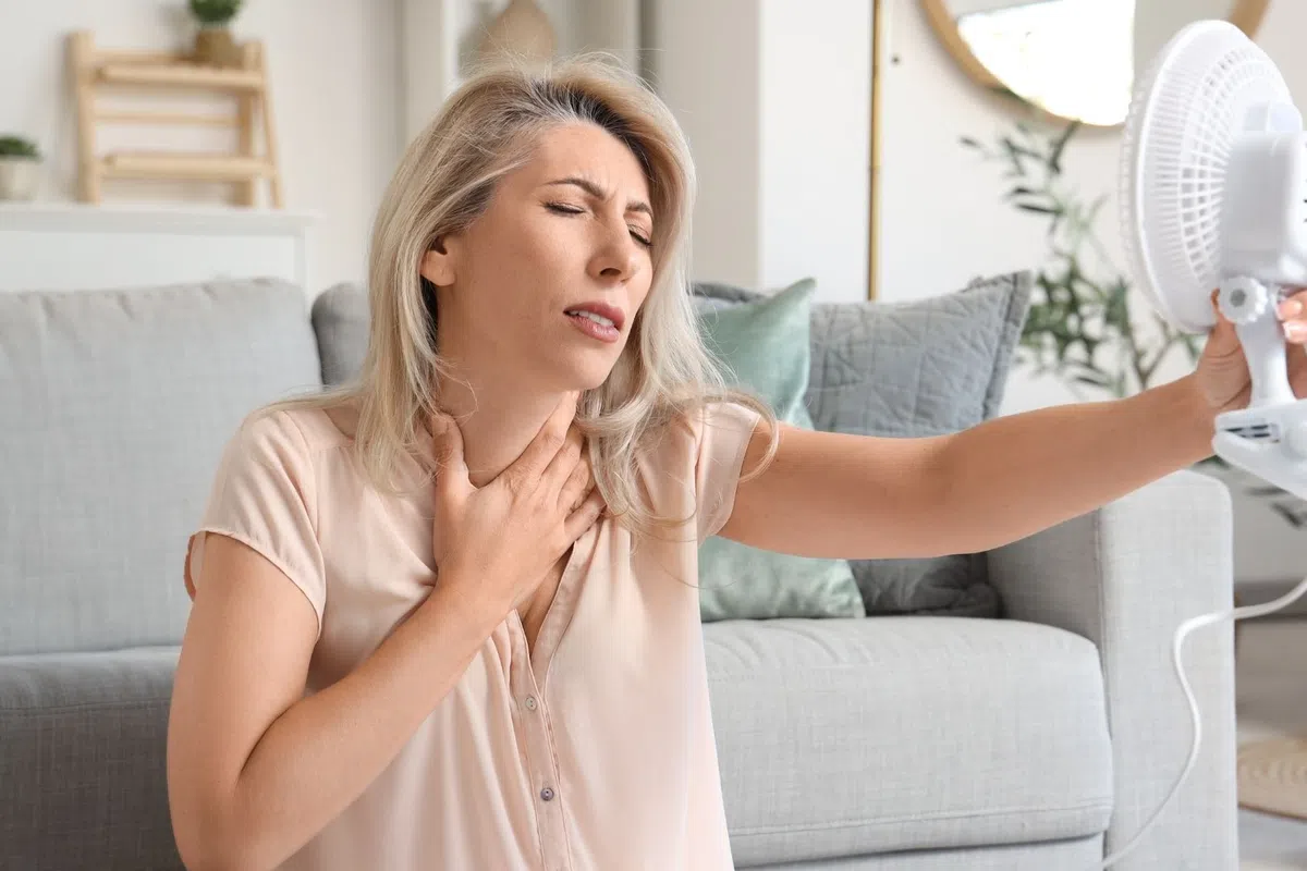 Woman sitting indoors feeling hot and uncomfortable while holding a small electric fan to cool down.