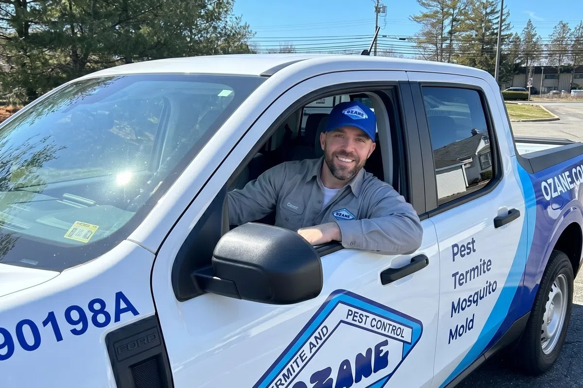 Smiling pest control worker sits in Ozane branded white and blue pickup truck on sunny day.