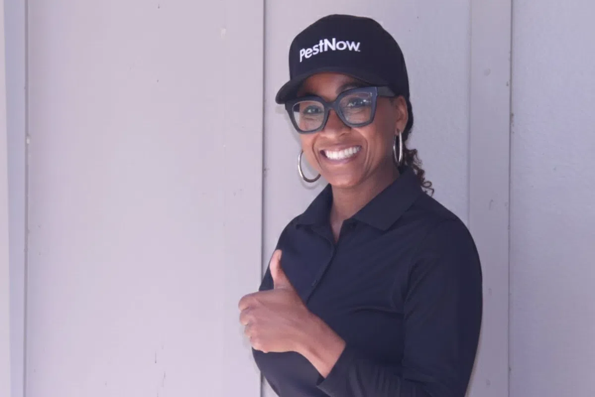 Smiling woman wearing PestNow cap and glasses giving thumbs up standing against a light grey wall