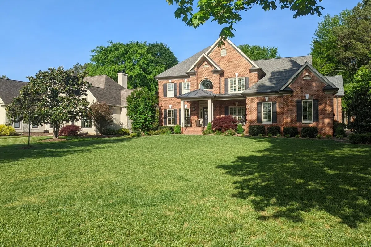 Two large houses with manicured lawns and trees under a clear blue sky in a suburban neighborhood