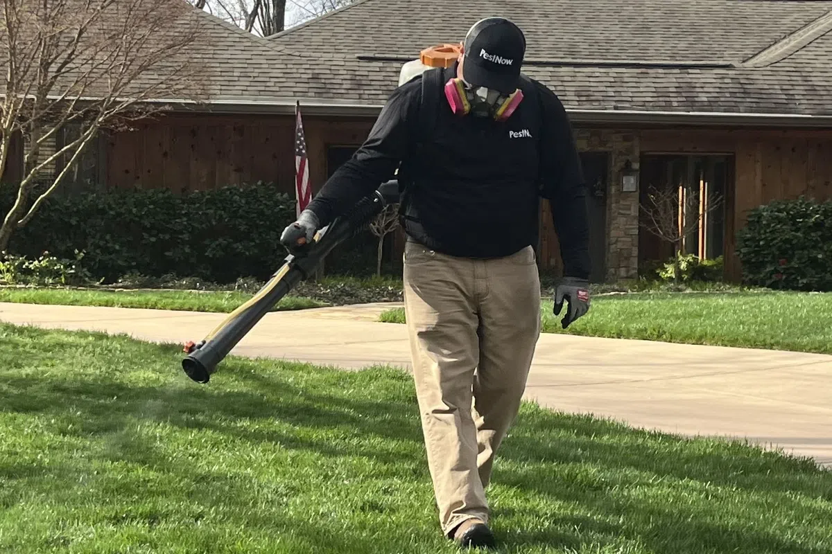 Pest control worker wearing protective gear using blower on green lawn in residential area