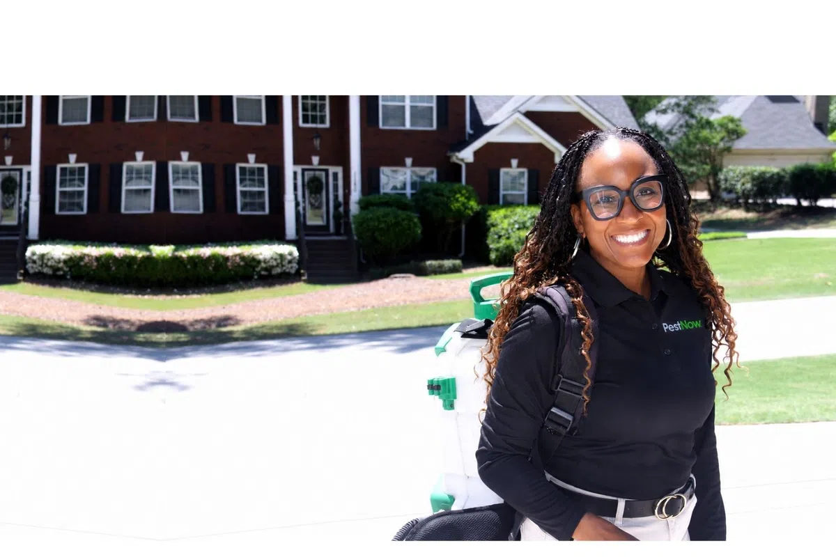 Smiling woman in PestNow uniform carrying pest control equipment in suburban neighborhood on sunny day