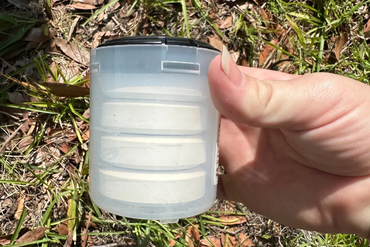 Hand holding a translucent geocache container outdoors on grass and fallen leaves in daylight.