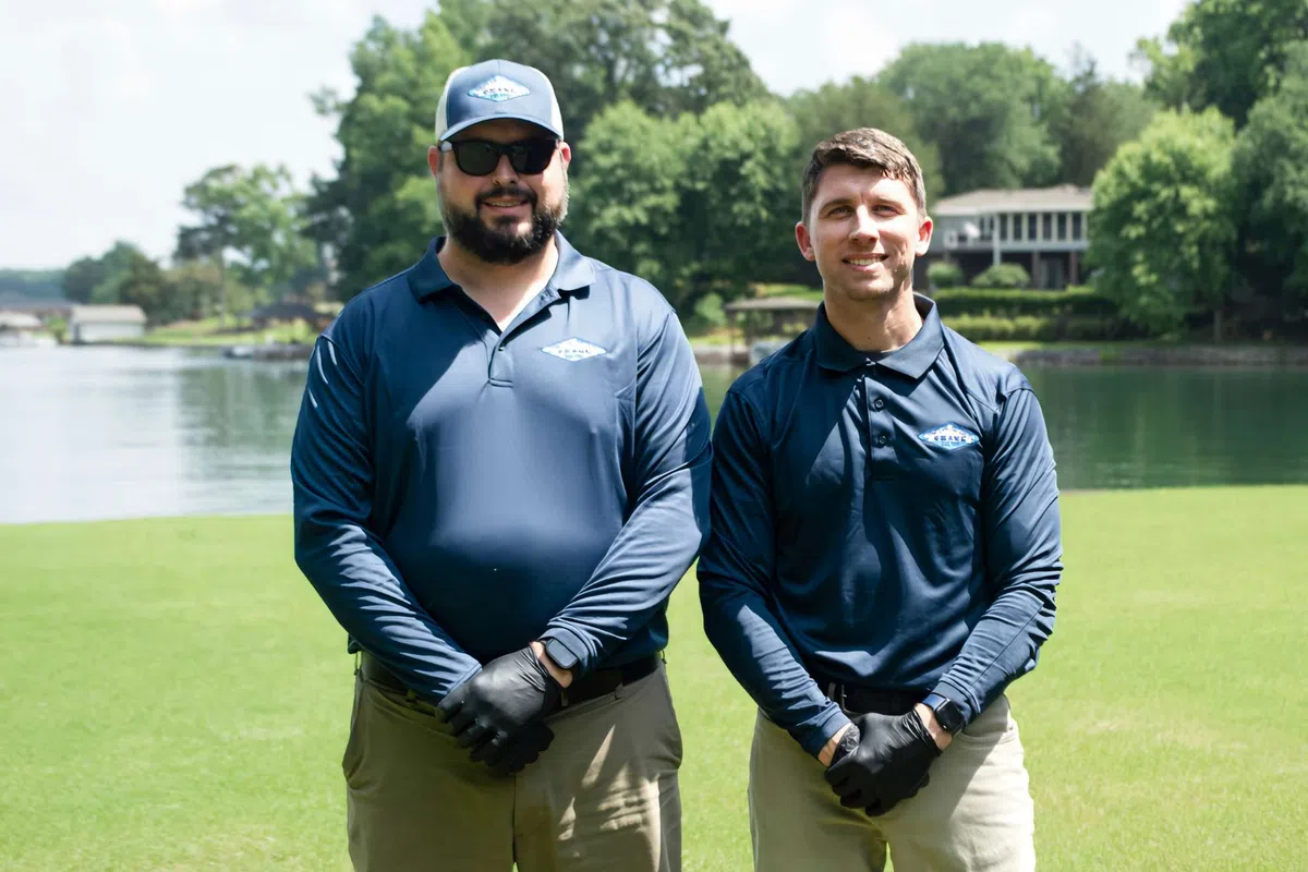 Two men in navy blue shirts and gloves standing on green grass by a lake with trees and houses in the background