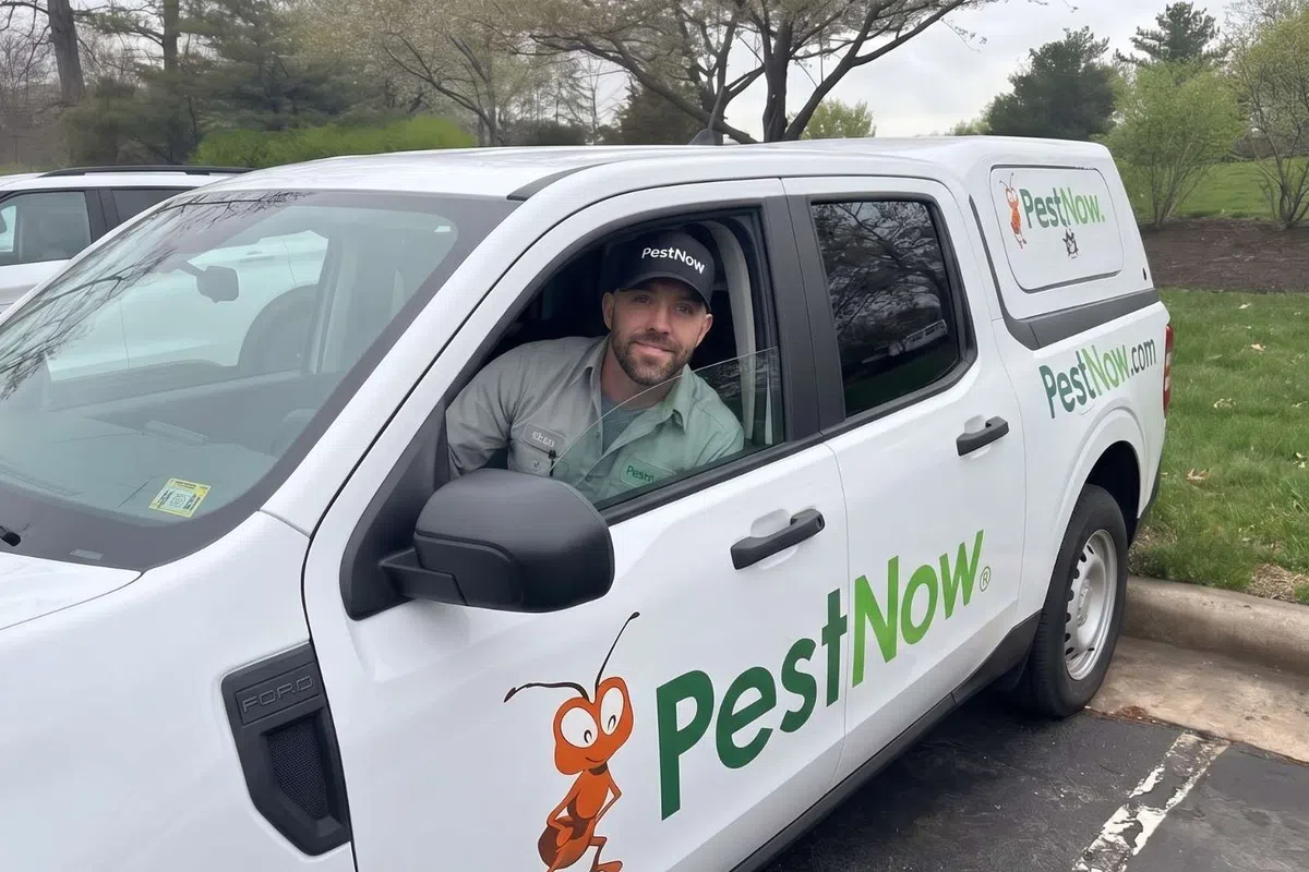 PestNow technician in uniform sitting inside branded white truck parked outdoors on a cloudy day.