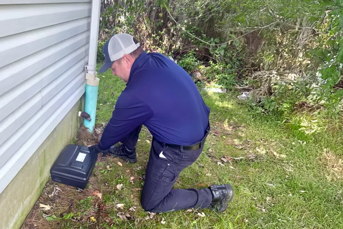 Technician kneeling outside a house inspecting or servicing a pest control bait station near the foundation.