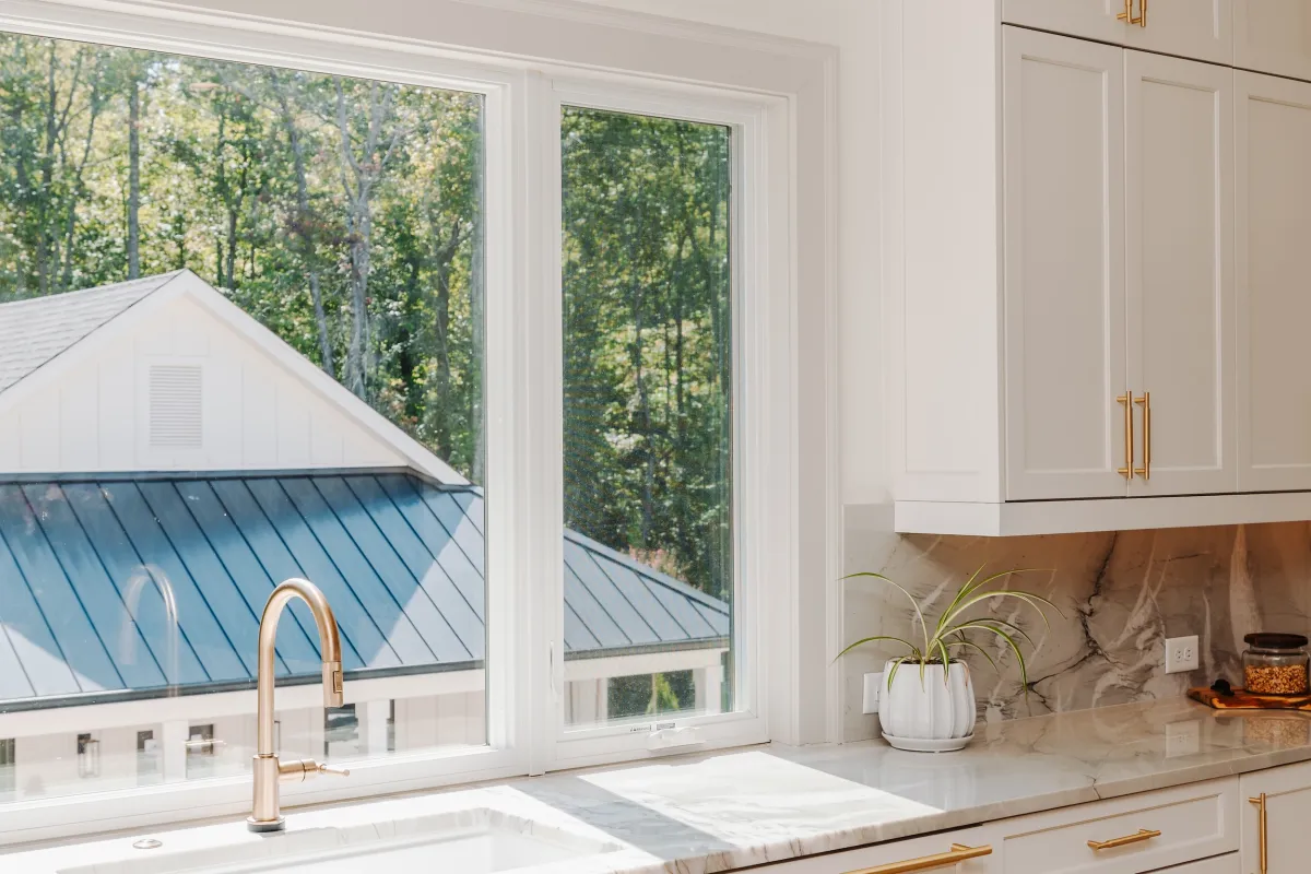 Bright white kitchen with gold hardware, marble countertops, large window, and green potted plant