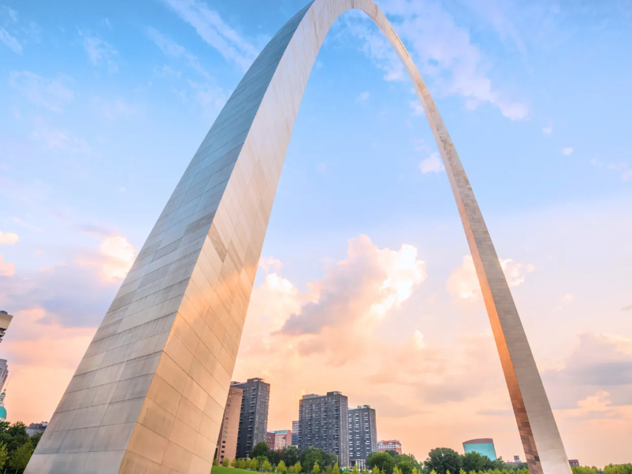 a large arch with grass and trees with Gateway Arch in the background