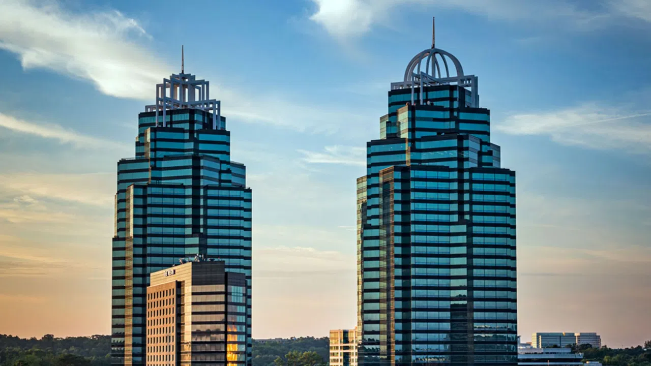 Twin modern glass skyscrapers against a blue sky during sunset with smaller buildings in the foreground.