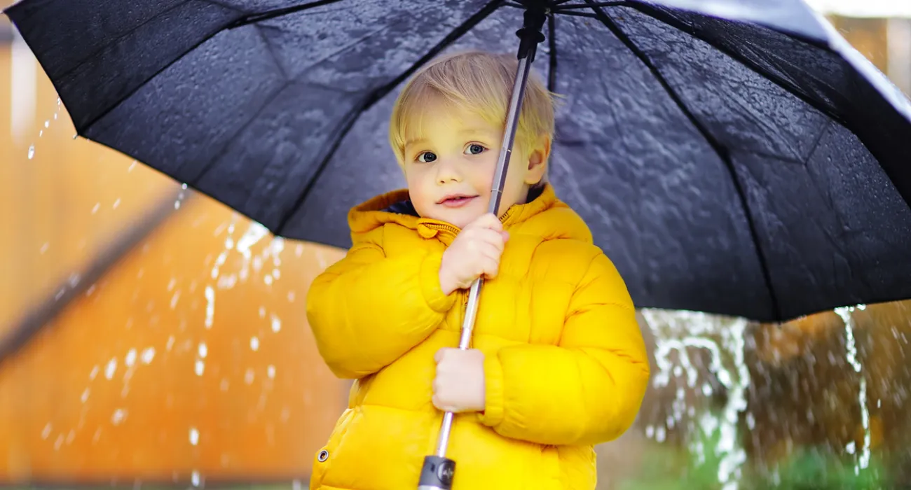Young child in a bright yellow jacket holding a black umbrella in the rain.