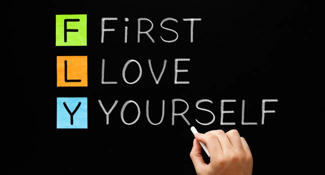 Hand writing the words FIRST LOVE YOURSELF with colorful blocks spelling FLY on blackboard.