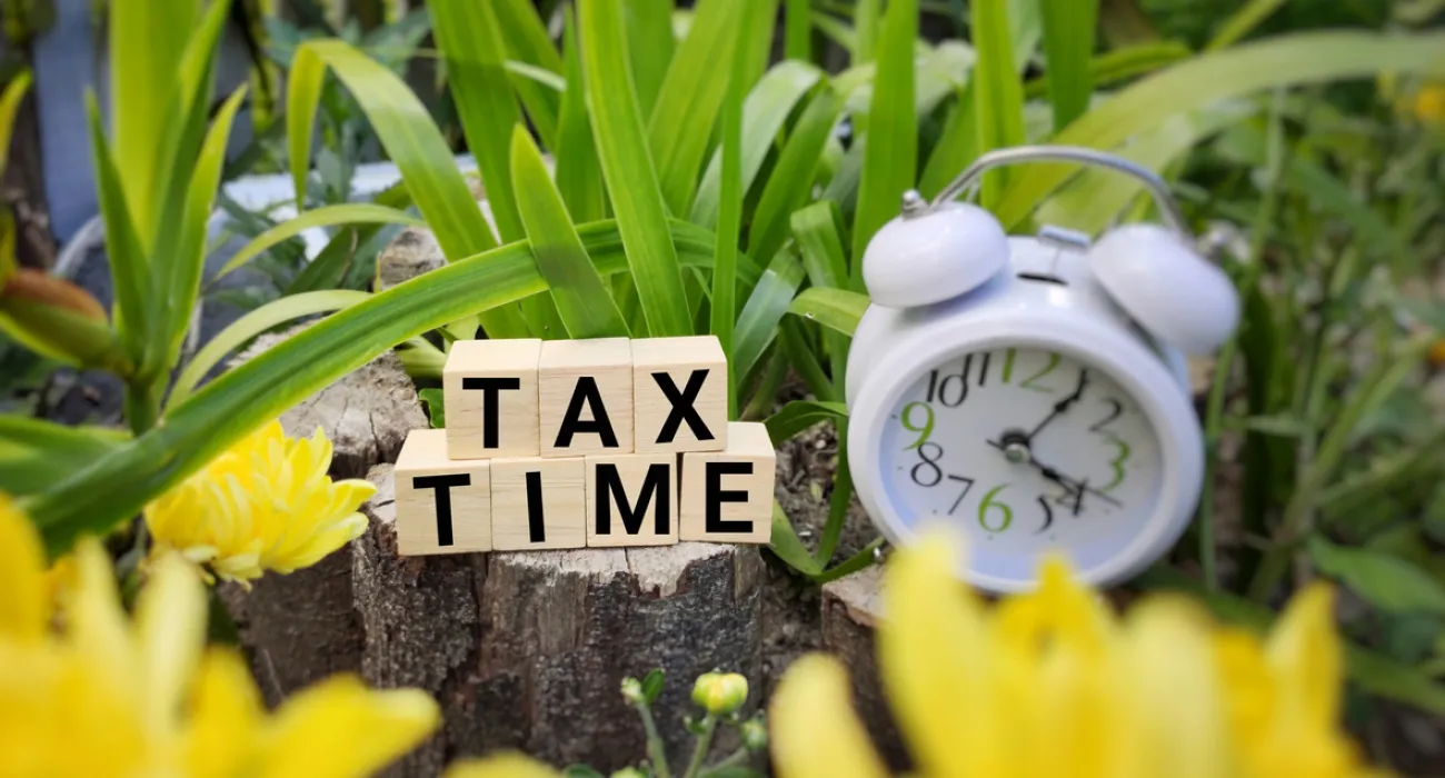 Blocks spell 'TAX TIME' next to a clock and vibrant yellow flowers in a garden setting.
