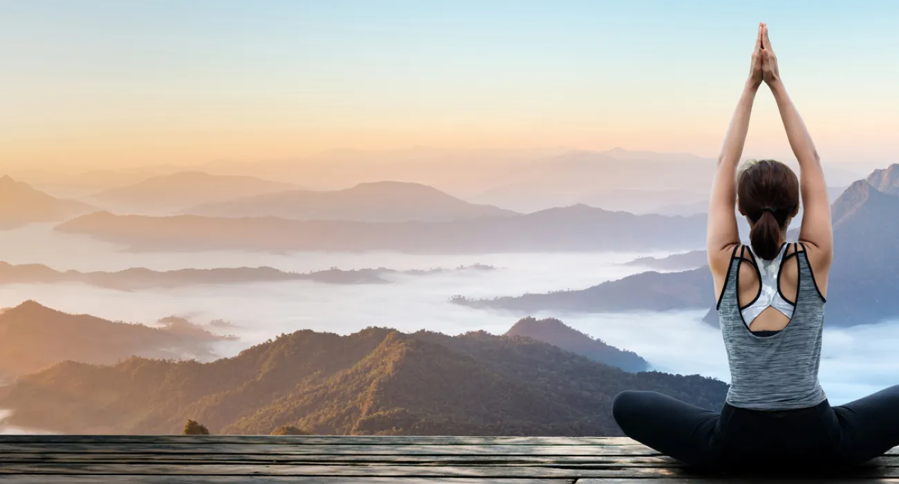 Woman practicing yoga on a wooden deck overlooking misty mountains at sunrise with clear sky.