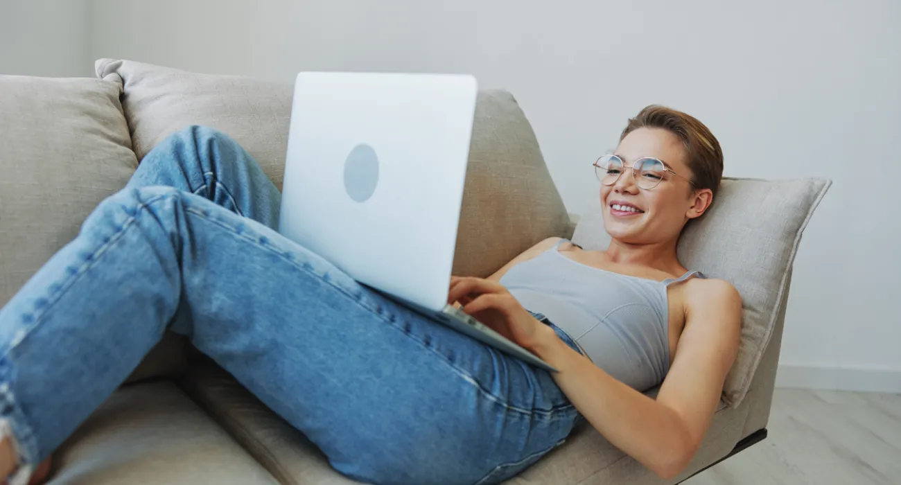 Young woman in glasses reclining on a couch with a laptop, smiling while working or browsing.